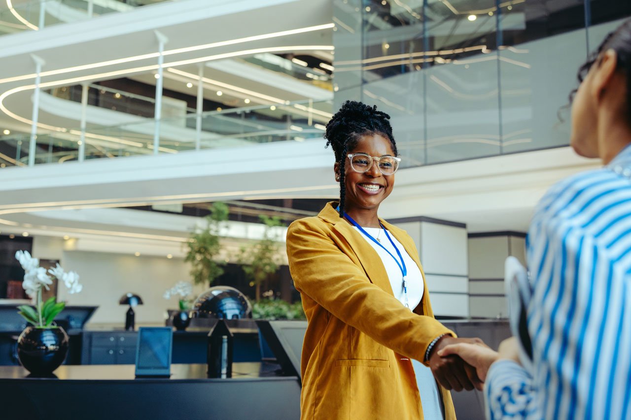 A woman wearing a yellow jacket is smiling and shaking hands with another individual during a successful meeting, in a modern, multi-level building decorated with plants and sleek, curved lighting.